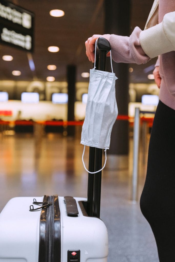 Person standing with luggage and face mask hanging, symbolizing pandemic travel.